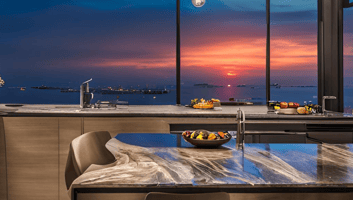 Through large windows, the vibrant colors of the sunset cast a warm glow over the scene, reflecting off the water, enhancing the serene and elegant atmosphere typical of upscale waterfront settings.